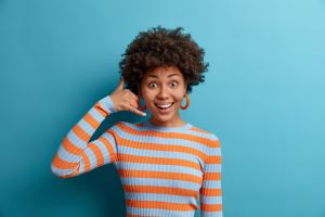 Smiling woman with curly hair standing against a teal background, wearing an orange and grey striped jumper and making a “call me” hand gesture near her ear.