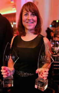 Smiling woman in a black dress holding two clear glass award trophies, standing at an awards event with warm lighting and a blurred background of other attendees.