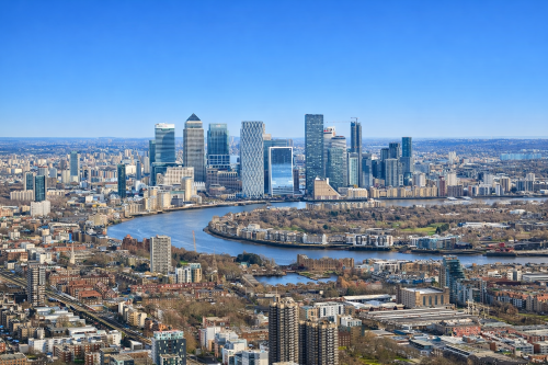 High resolution aerial view of Canary Wharf and the River Thames in London under a clear blue sky, showing modern skyscrapers, surrounding residential buildings, green spaces, and railway lines in sharp detail.