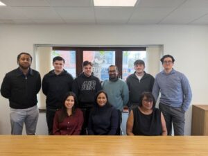 Case Managers group photo of eight colleagues in an office, with five people standing and three seated behind a wooden desk, posing in front of a window under bright ceiling lights.